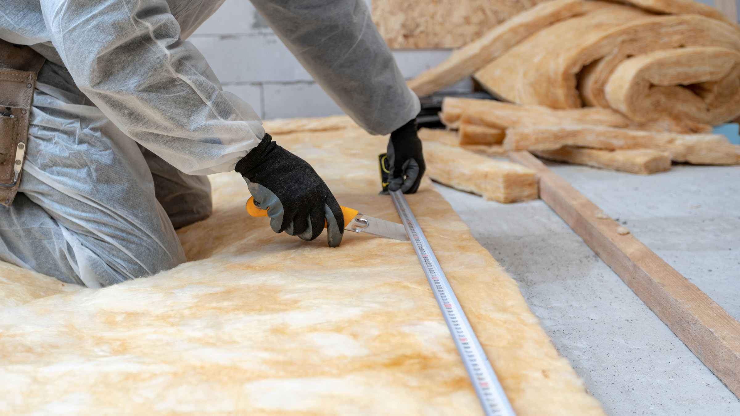Cropped view of professional workman in protective workwear using knife and holding measuring tape over material, installing thermal insulation rock wool for basement